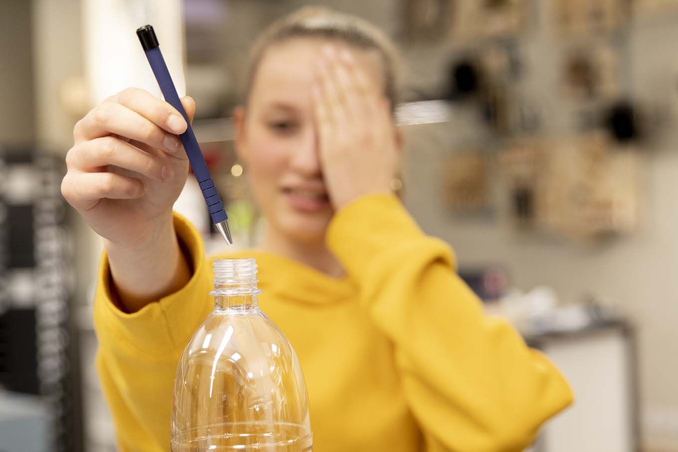 Jente holder venstre hånd foran venstre øye mens hun holder en penn i høyre hånd og fører pennen ned i en plastflaske. Foto